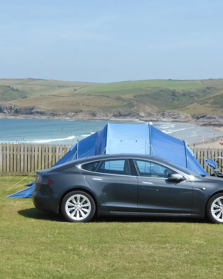 Tesla Model S neben einem Zelt auf einem Campingplatz an der Küste, im Hintergrund Bucht und Sandstrand